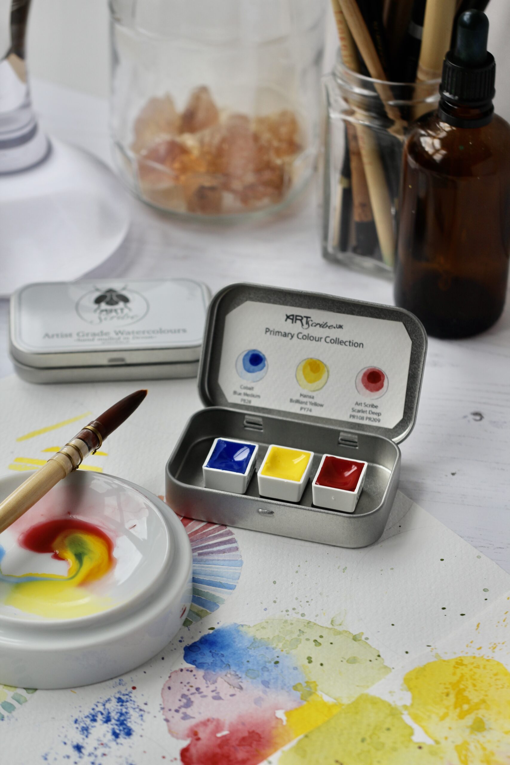 open tin of Handmade watercolour paint by Art Scribe Primary Collection. A paintbrush, palette with watercolour paint and paper with splashes of colour from the tin sit on the table. Jars and bottles are in the background.