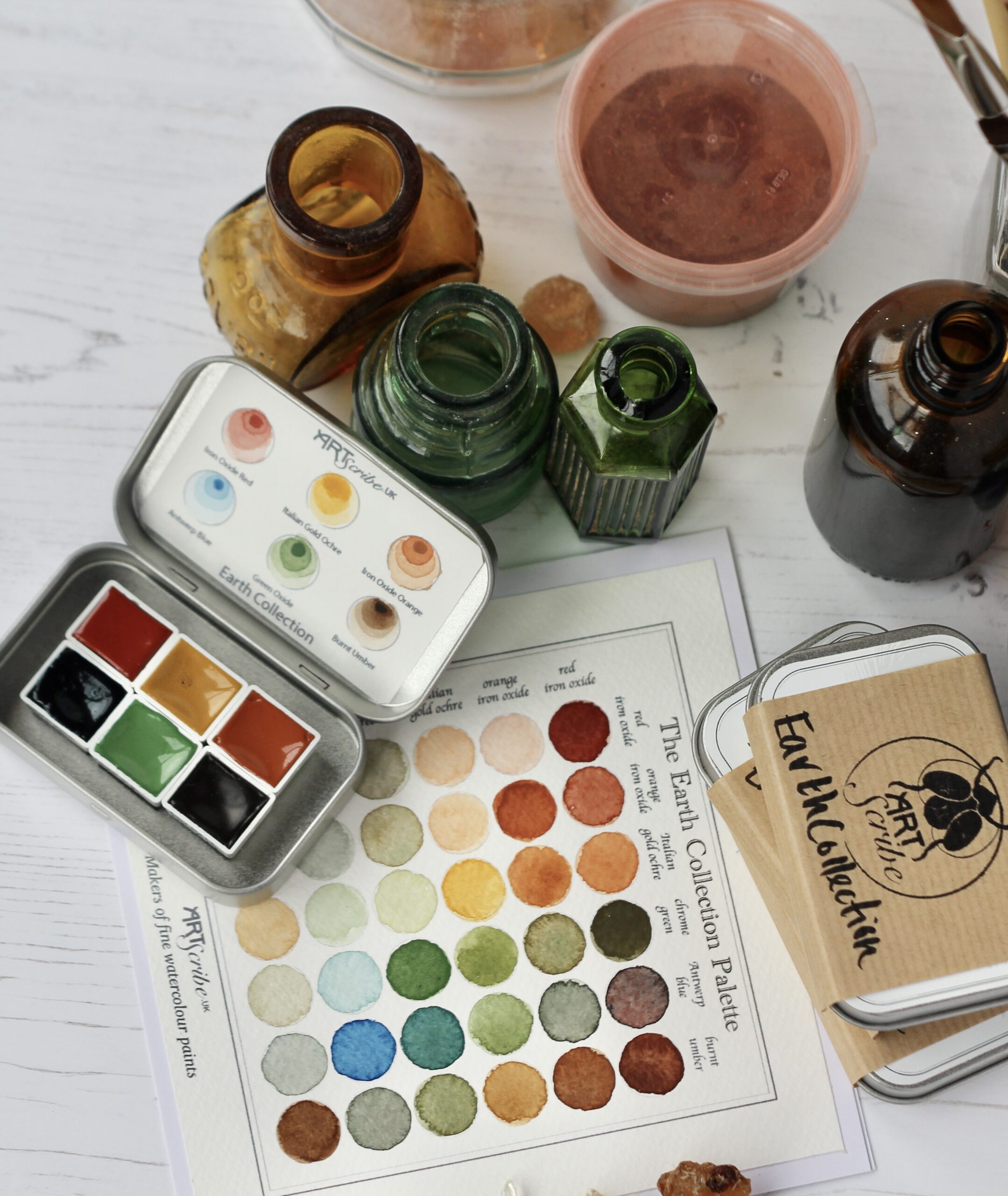 An overview of Art Scribe Earth Collection Handmade watercolour paint tin opened. it sits upon a colour mixing chart with old bottles and tubs of pigment.