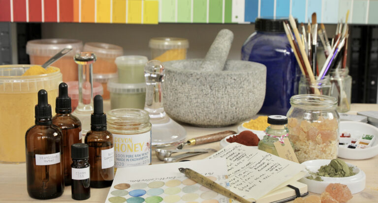 A table full of paint making tools and ingredients. There are brown bottles with pipettes, tubs of coloured pigments, a pestle and mortar, paint brushes, measuring spoons and an open book with handwritten paint making recipe.