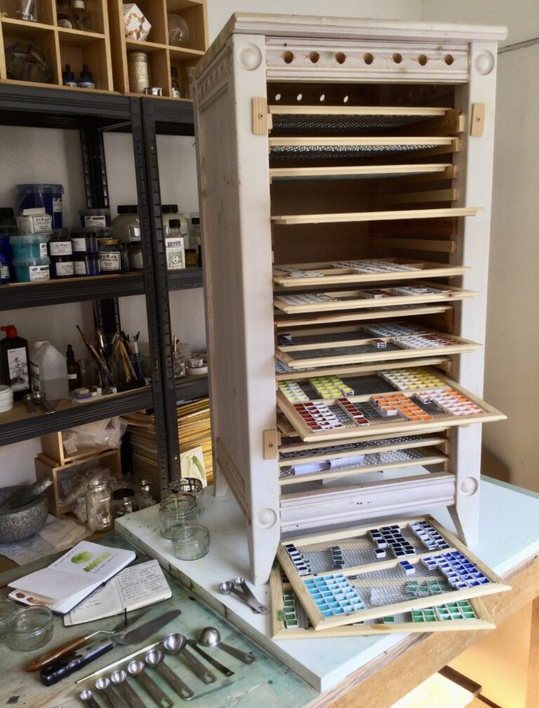 Image shows number three of three favourite paint making tools. A quirky looking cabinet inside a paint making studio stands on top of a bench. the open front shows many flat draws containing hand made paint.