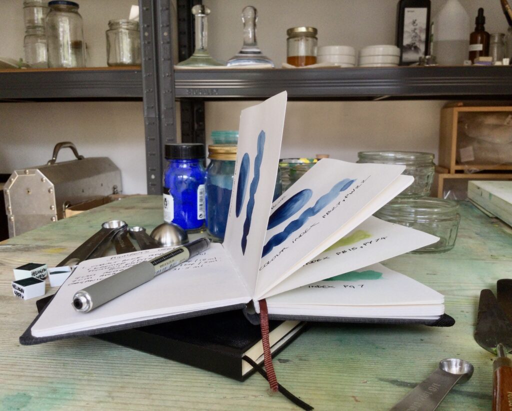 Image shows second of three favourite paint making tools. A small black book lays open on a bench in an art studio.